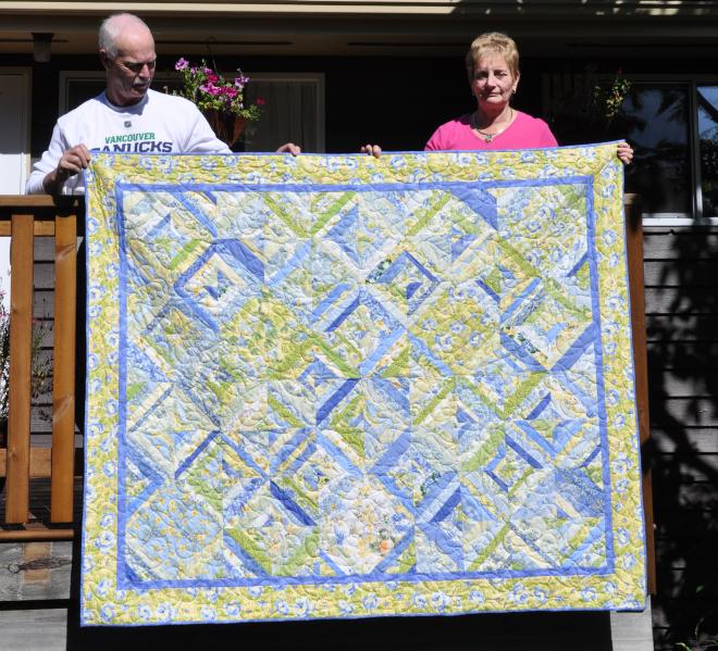 StrataVarious, a geometric patchwork quilt in yellows, greens and blues, lap size, being held up by Barbara and Bill.